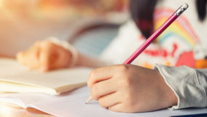 Girl sits at desk doing homework, hand writing in notebook