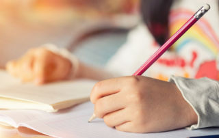 Girl sits at desk doing homework, hand writing in notebook